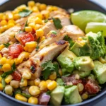A black bowl filled with grilled chicken, corn, avocado, diced tomatoes, red onion, cilantro, and lime wedges.