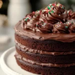 A three-layer chocolate cake with chocolate frosting and sprinkles sits on a white cake stand.