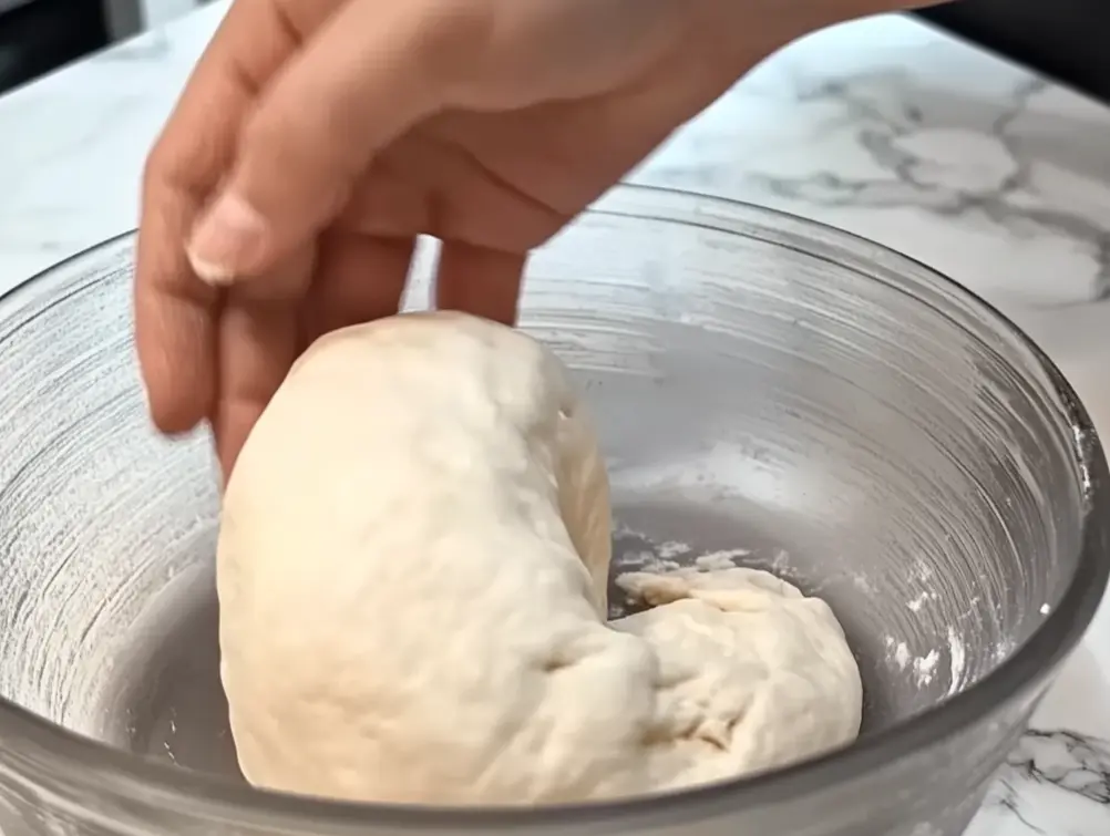 Hand kneading a soft dough ball in a glass bowl on a marble countertop.