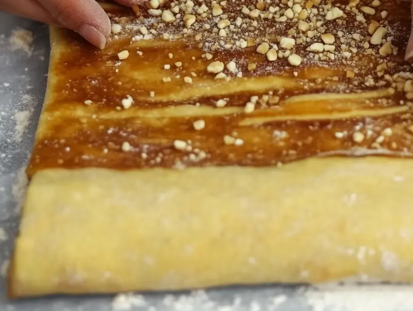 A person folds a sheet of dough topped with chopped nuts and cinnamon.
