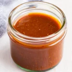 A small, clear glass jar filled with a vibrant, reddish-brown sauce.